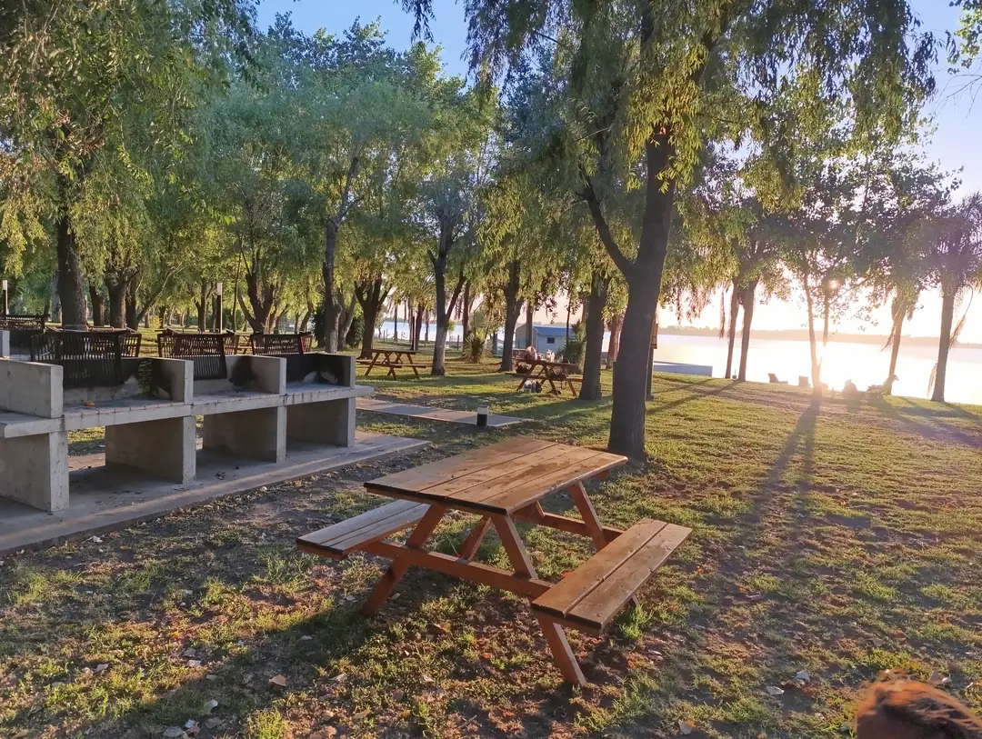 Mesas de madera y parrillas bajo los árboles, con el sol bajo sobre la laguna.