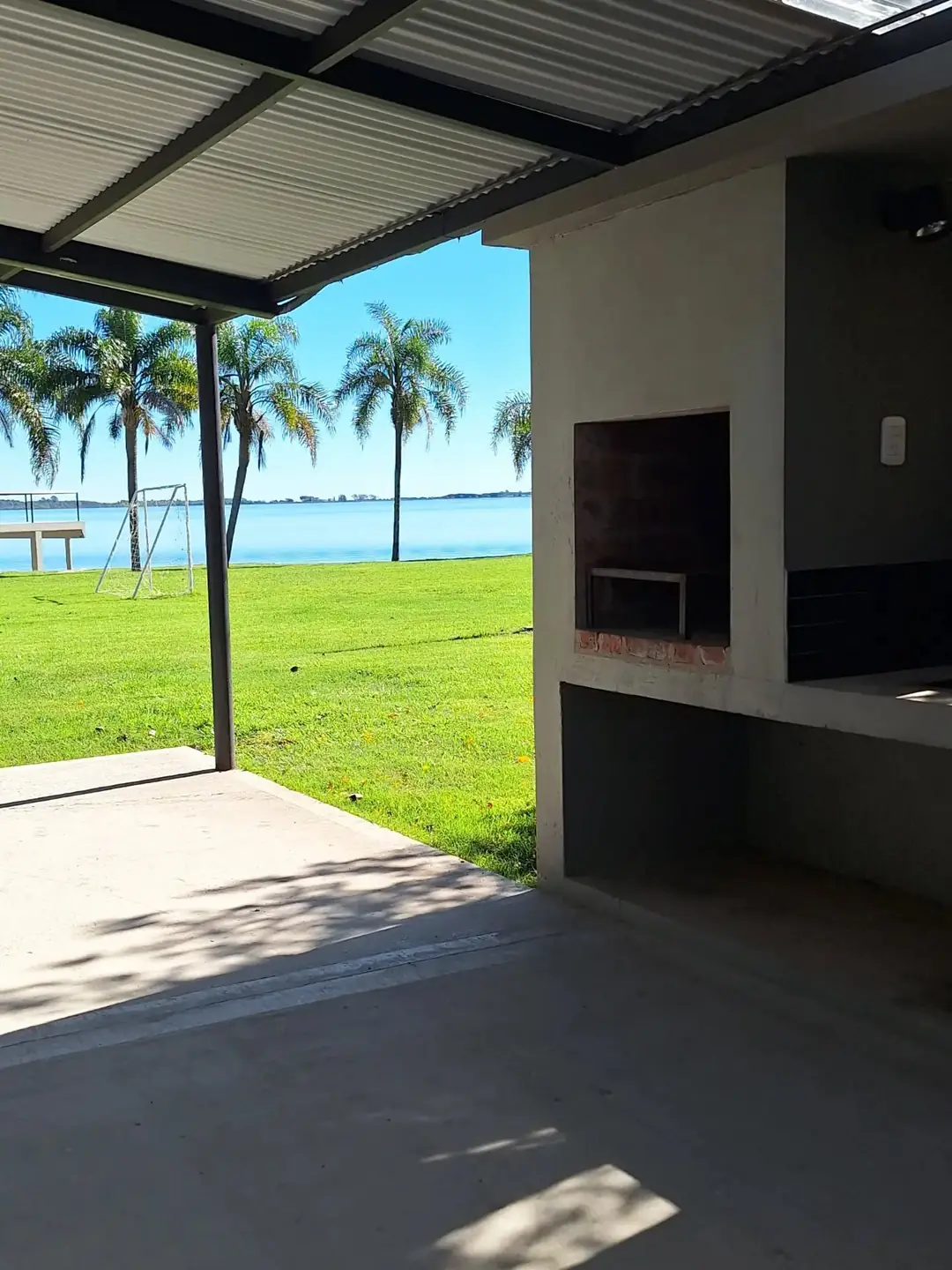 La galería techada del monoambiente con su parrilla, y la laguna con palmeras de fondo.