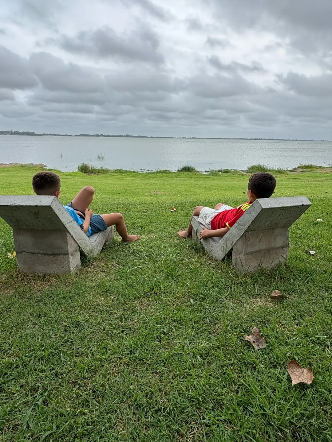Dos chicos en reposeras de hormigón mirando la laguna.